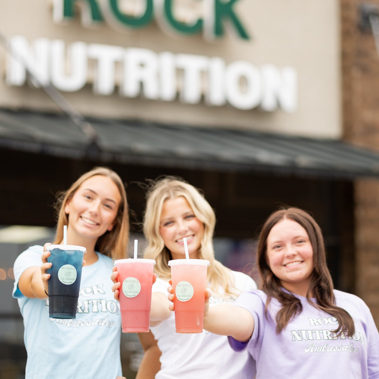 Three Rock Nutrition team members holding loaded teas in front of the Owasso storefront sign